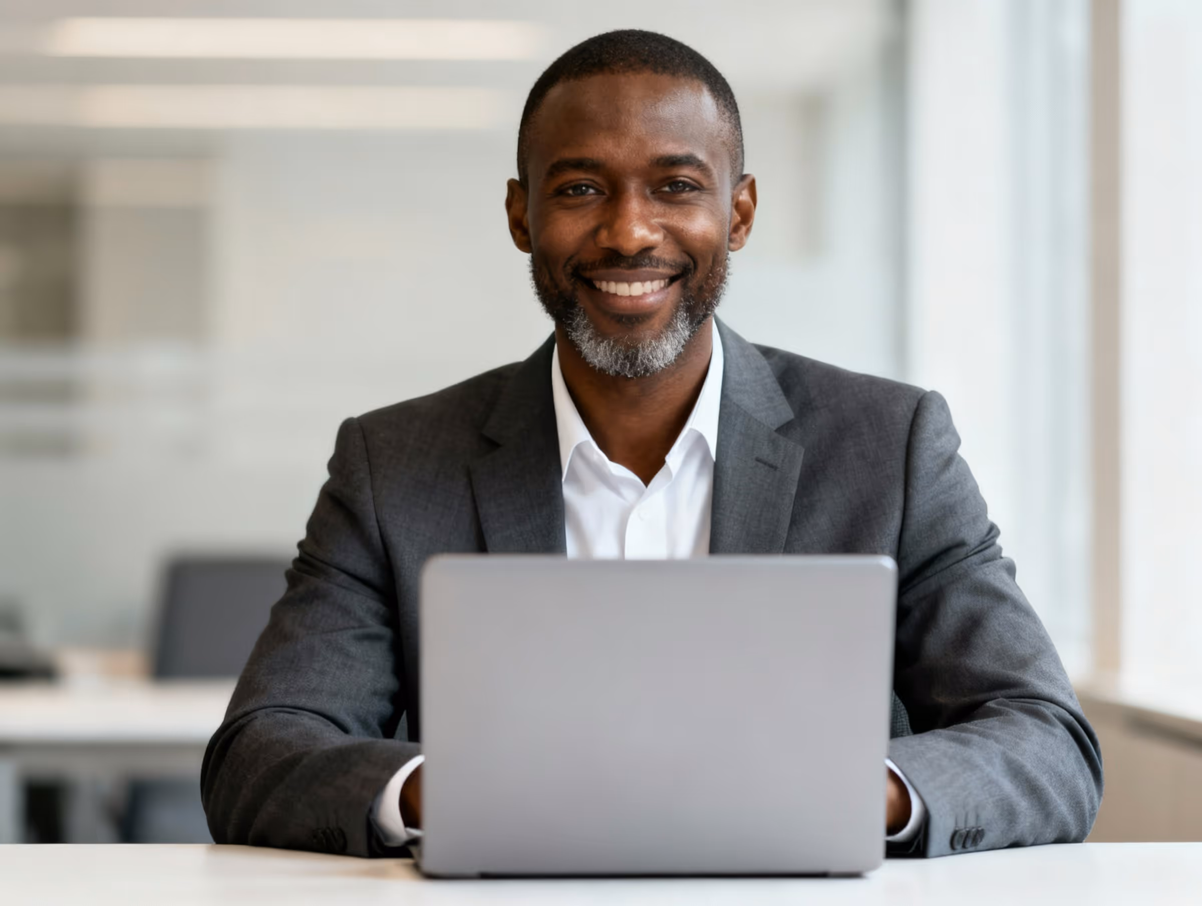 Smiling man in a suit sitting at a desk with an open laptop in an office.