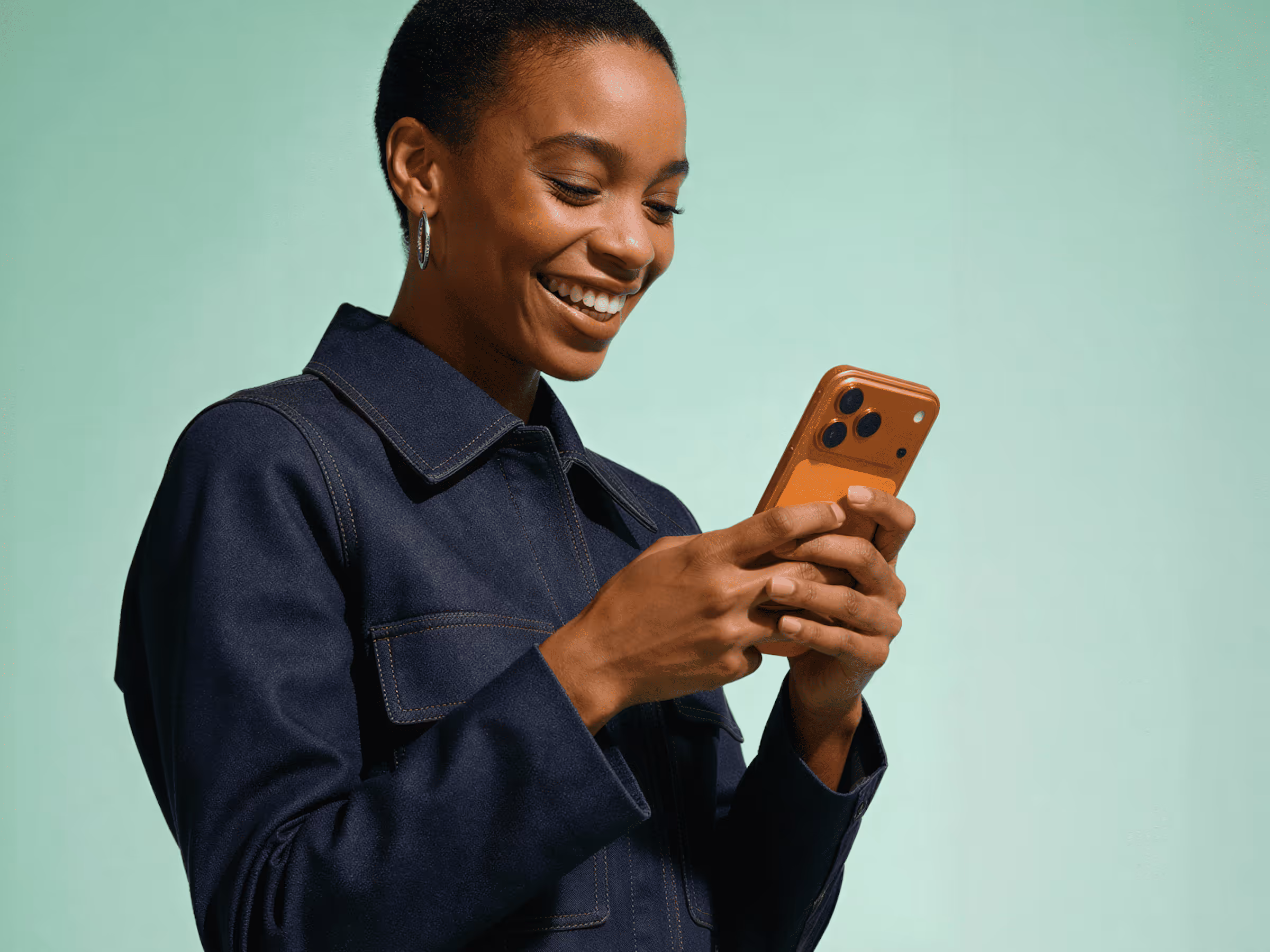 Smiling woman in dark jacket looking at a smartphone with a brown case against a light green background.