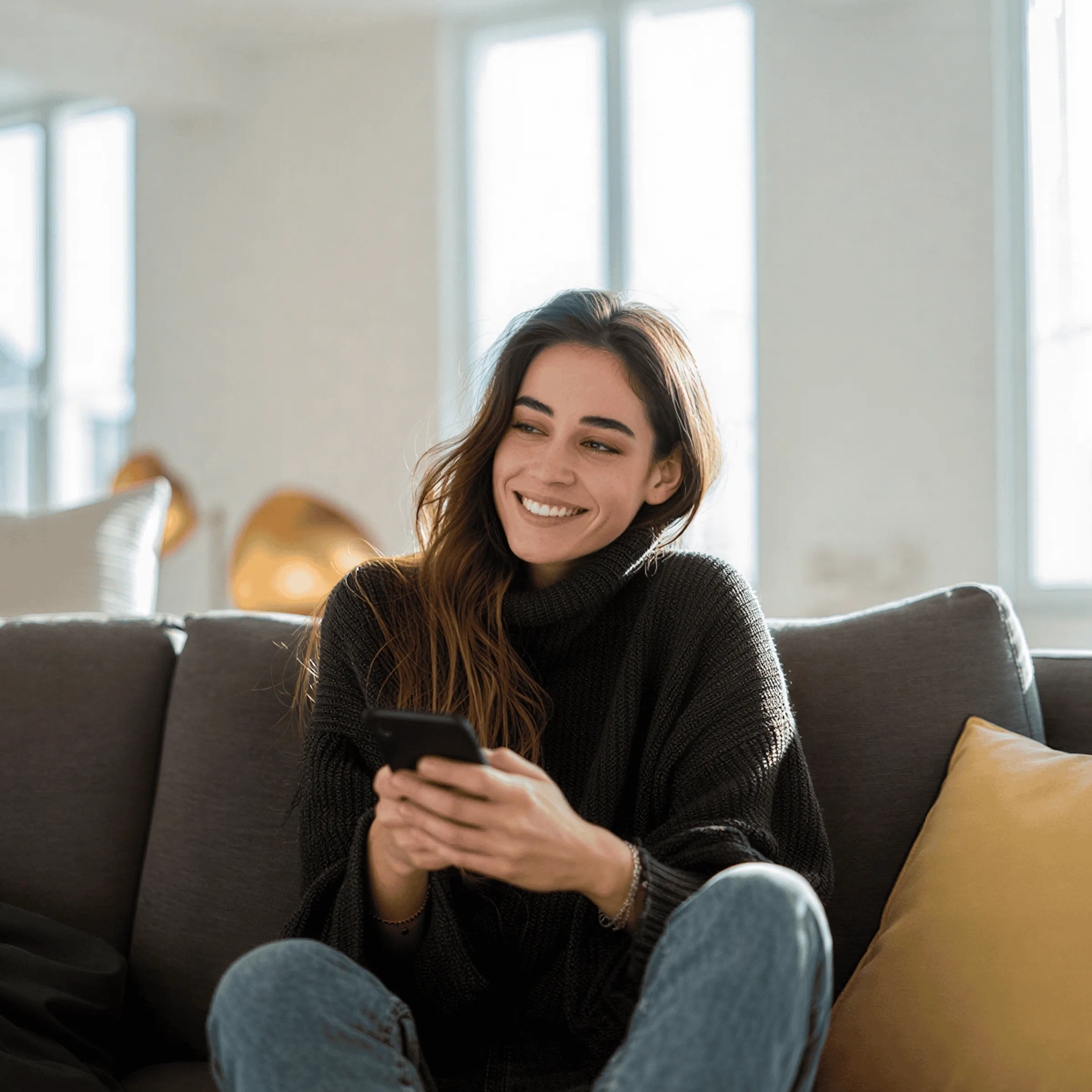 Smiling woman in a black sweater sitting on a couch holding a smartphone indoors.