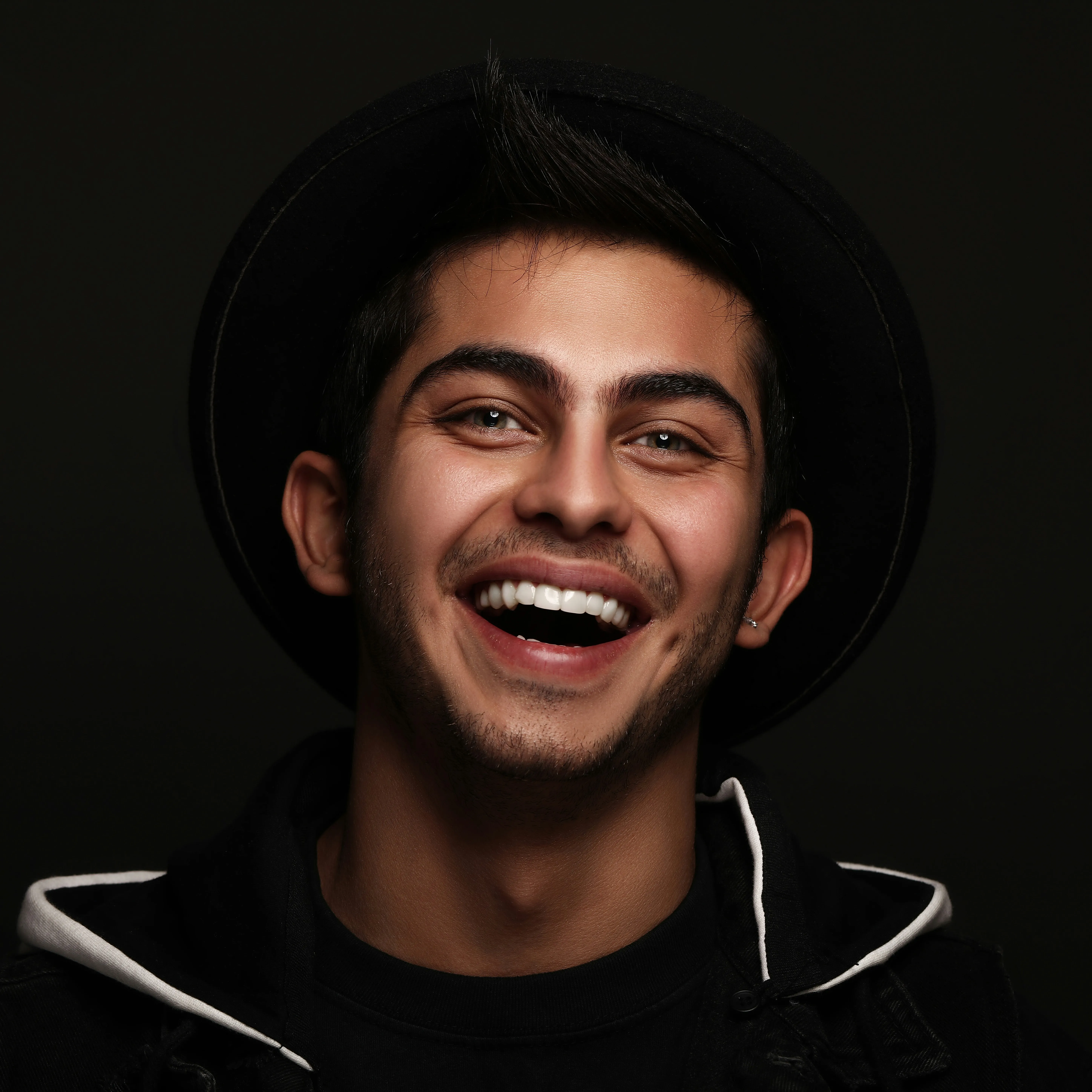 Young man with a black hat smiling broadly against a dark background.