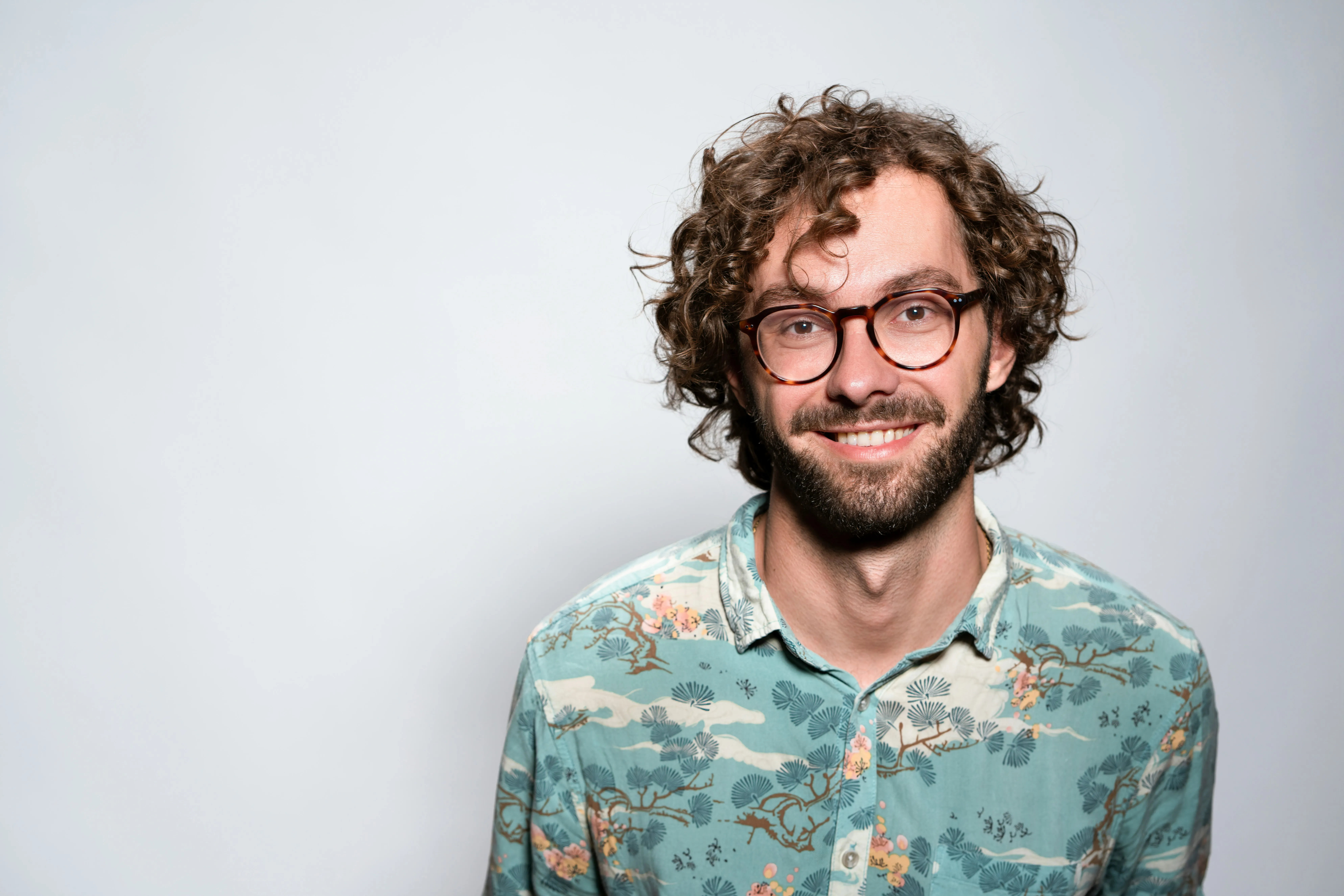 Smiling young man with curly hair, beard, glasses, and floral patterned shirt against plain background.