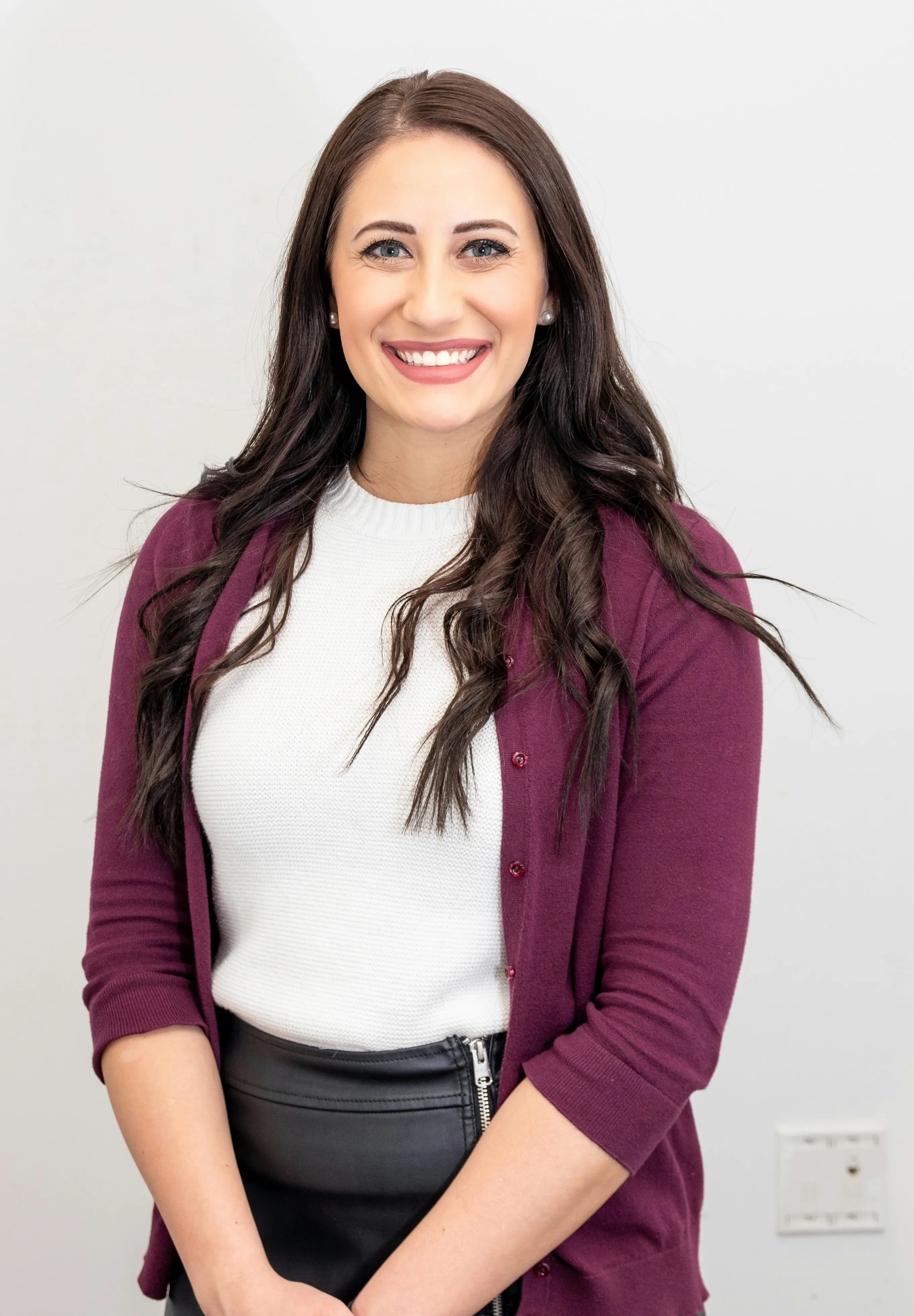Smiling woman with long dark hair wearing a white top and purple cardigan standing in front of a white wall.