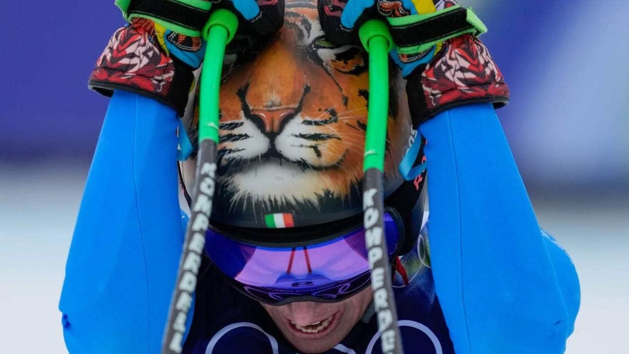 Primo piano di Federica Brignone con casco decorato con il muso di una tigre e bandiera italiana, occhiali da sci a specchio viola e bastoncini verdi sollevati sopra la testa, in un momento di intensa concentrazione durante la gara.