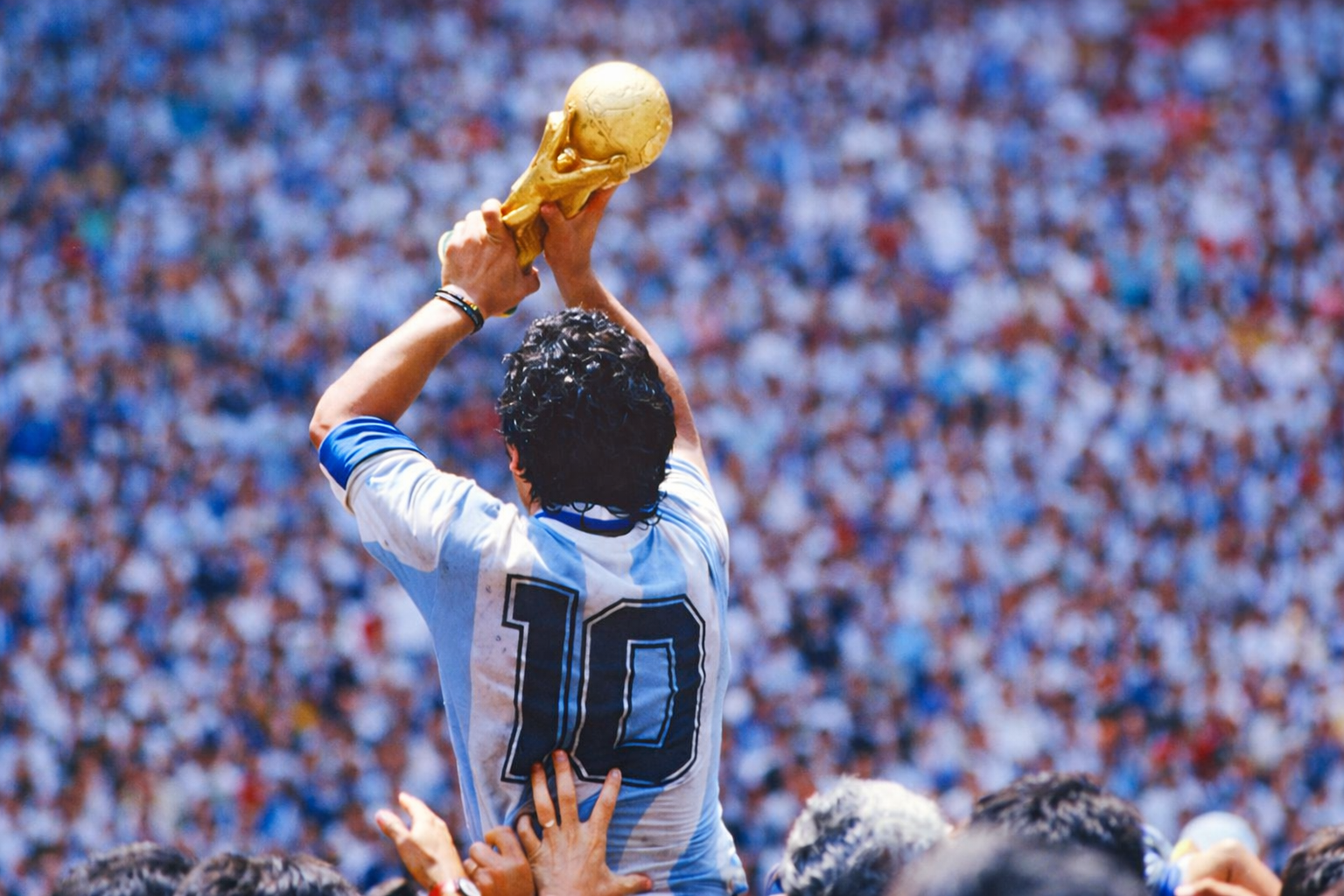 Diego Maradona solleva la Coppa del Mondo indossando la maglia numero 10 dell'Argentina mentre viene portato in trionfo dai tifosi in uno stadio gremito.