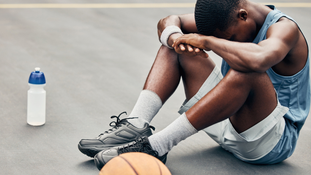 Un ragazzo seduto su un campo da basket all’aperto, con la testa appoggiata sulle braccia in segno di stanchezza o frustrazione. Accanto a lui ci sono una palla da basket in primo piano e una borraccia d’acqua. Indossa abbigliamento sportivo e sembra aver appena finito di allenarsi. 