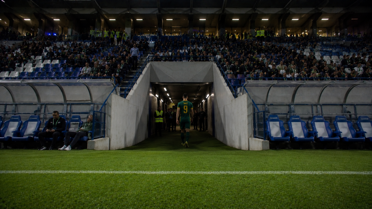 Un calciatore solitario con la maglia verde numero 9 entra nel tunnel degli spogliatoi di uno stadio moderno, ripreso dal campo in un’atmosfera cinematografica notturna. Gli spalti pieni e le luci intense dello stadio contrastano con il tunnel scuro e realistico sullo sfondo. 