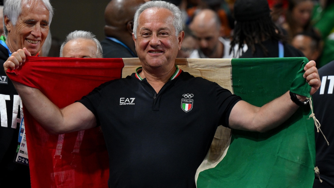 Velasco sorride al centro della foto mentre tiene aperta una grande bandiera italiana, circondato da membri dello staff e pubblico sullo sfondo. Indossa una polo nera con lo stemma dell’Italia e il simbolo olimpico. 