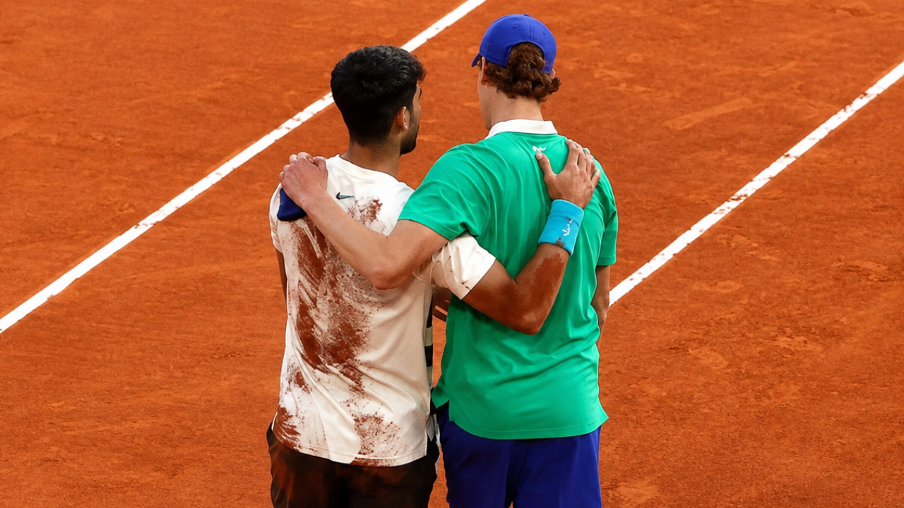 Sinner e Alcaraz camminano abbracciati su un campo in terra rossa, in un gesto di amicizia e sportività al termine della partita. 