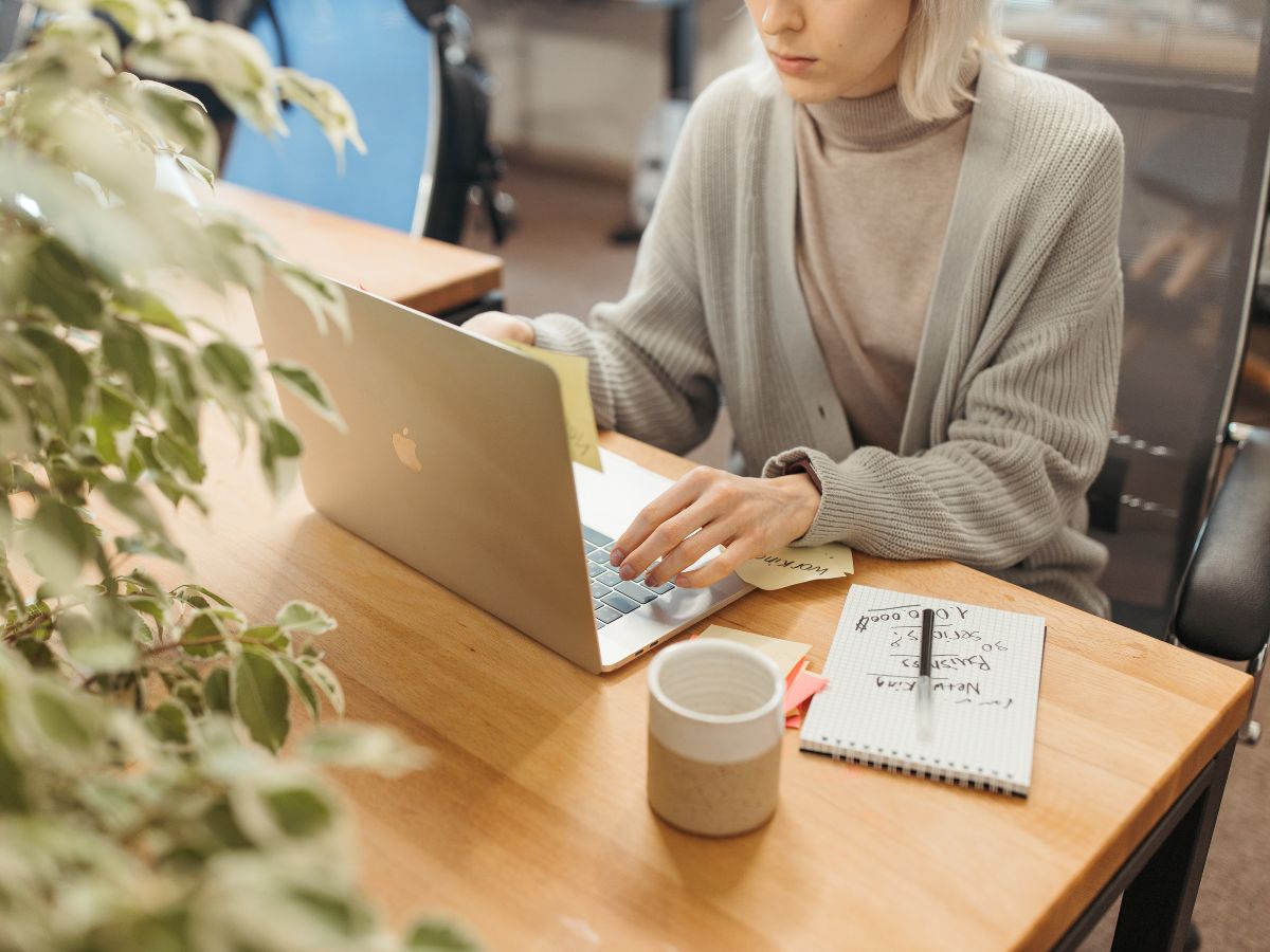 A person with bleach blonde hair, looking at a silver Macbook screen, comparing notes on a post it to what she sees on the screen.
