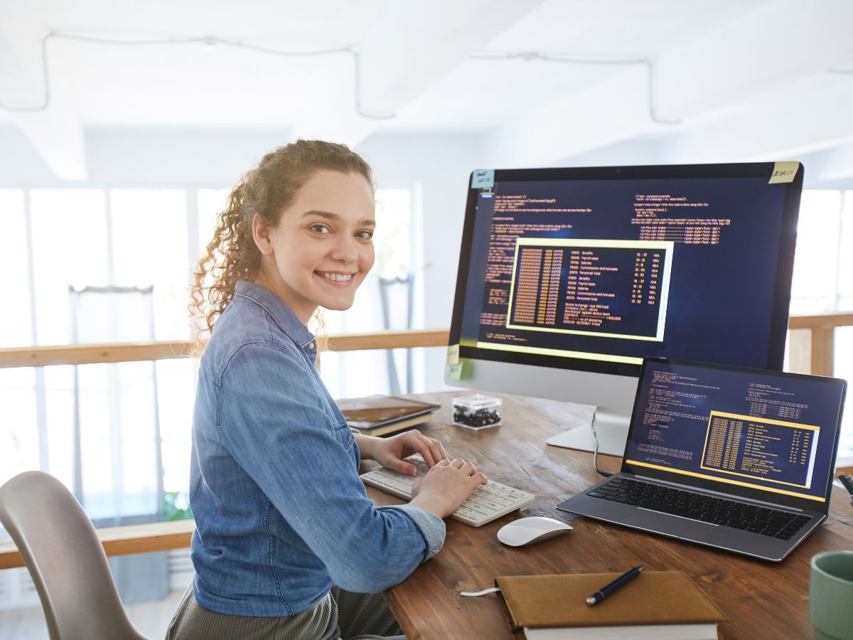 A woman smiling while sitting in front of a desktop computer displaying codes in it.