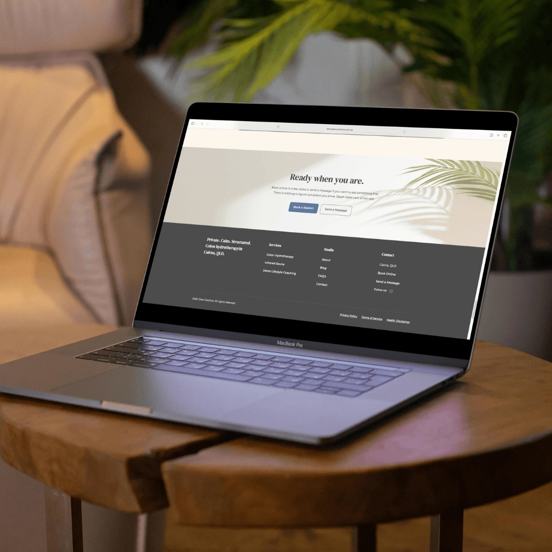 Laptop mockup on a wooden side table displaying the "Ready when you are" call-to-action and footer section of the Glow Colonics website