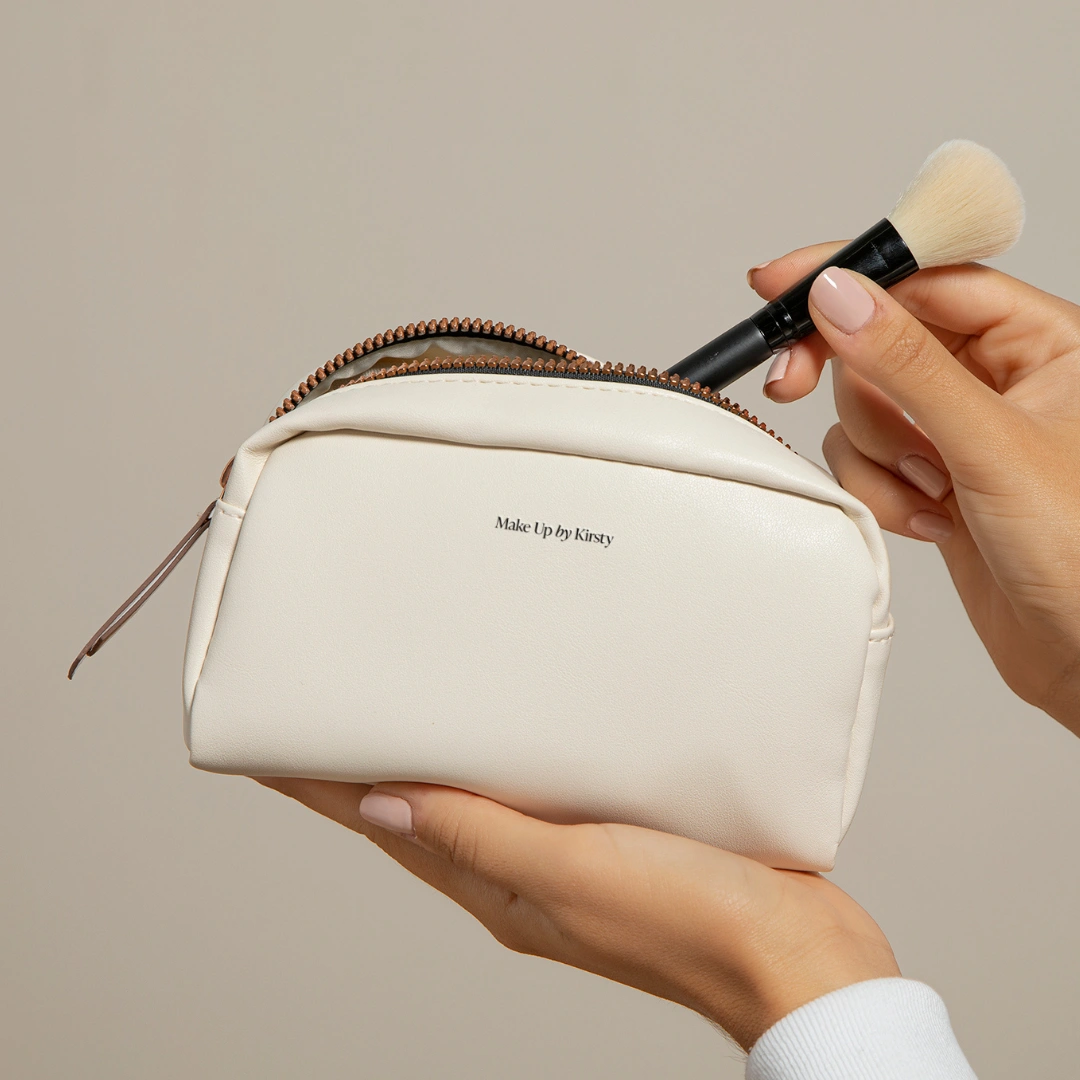 Hands holding a cream branded Make Up by Kirsty cosmetics pouch while inserting a makeup brush