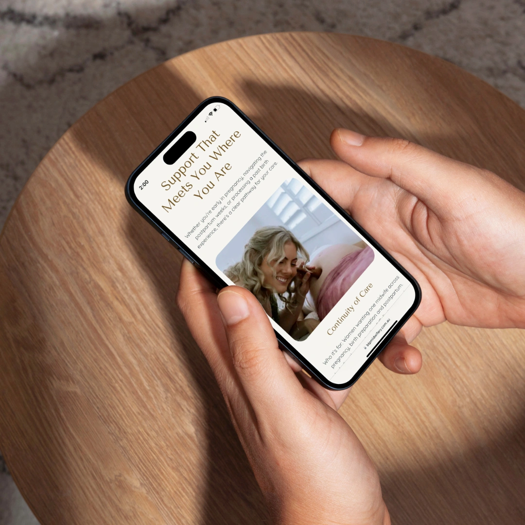 Hands holding a smartphone on a wooden side table, displaying the "Support That Meets You Where You Are" Continuity of Care page on the Beneath the Palms Midwifery website