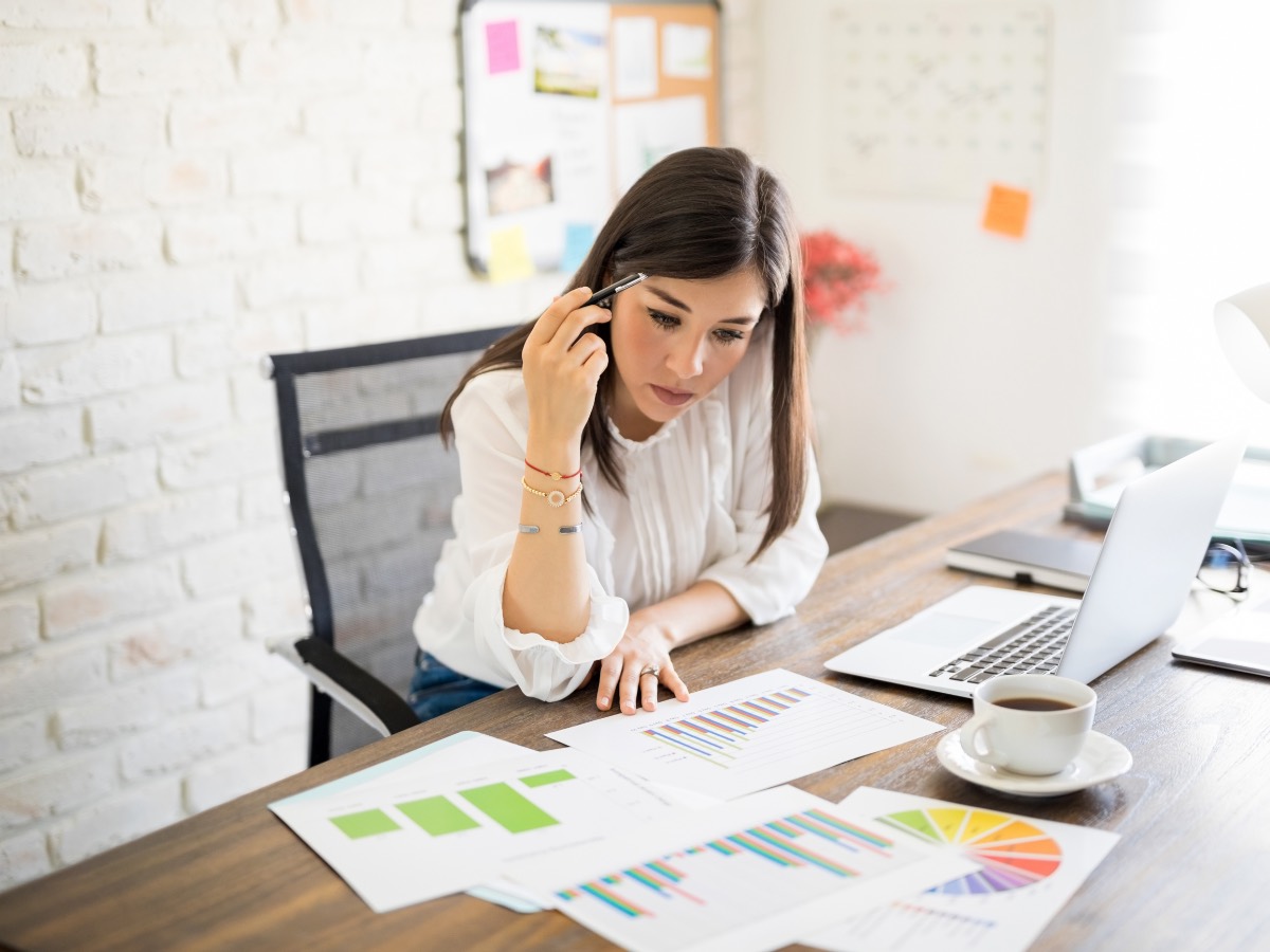 Concerned woman reviewing printed charts and reports at a desk with a laptop and coffee
