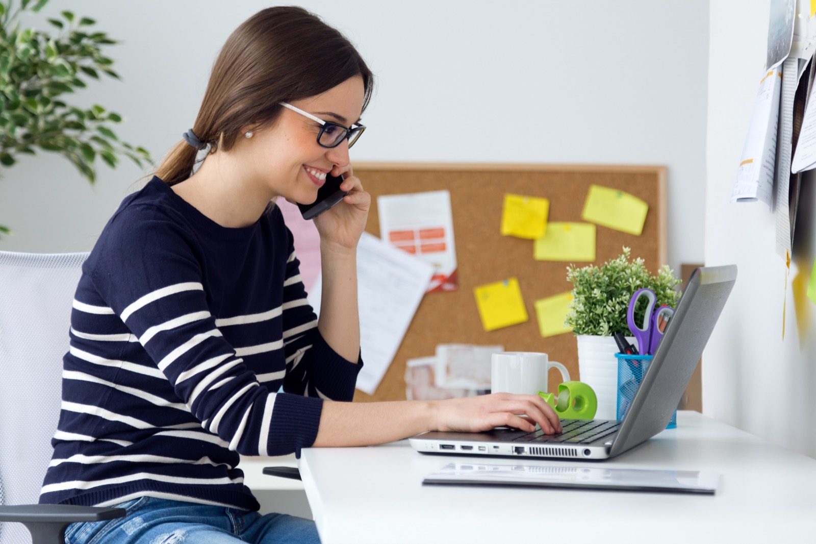 Smiling woman on the phone while typing on a laptop at a desk