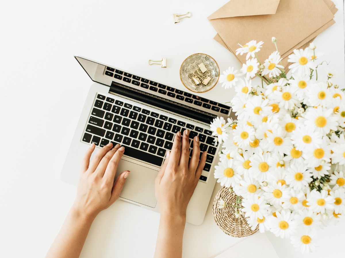Overhead view of hands typing on a laptop beside a bunch of daisies and envelopes