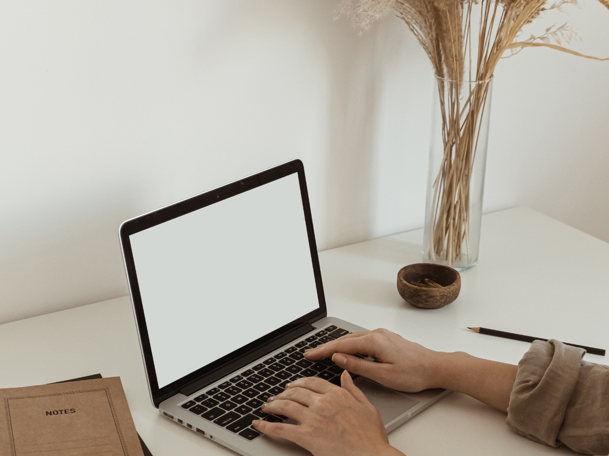 Hands typing on a laptop with a blank screen next to a vase of dried grasses