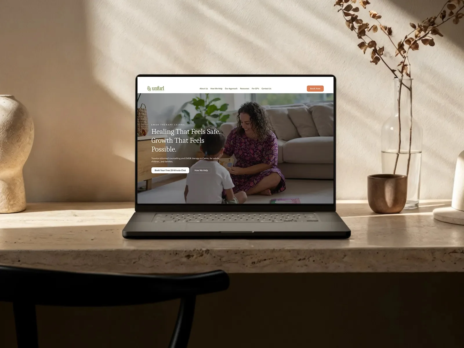 Laptop on a stone side table showing the Unfurl homepage, 'Healing That Feels Safe. Growth That Feels Possible.'