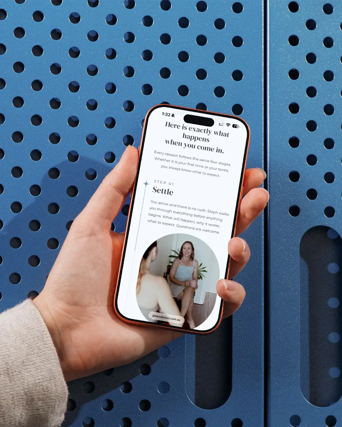Hand holding a phone against a blue wall showing the Glow Colonics page 'Here is exactly what happens when you come in.'