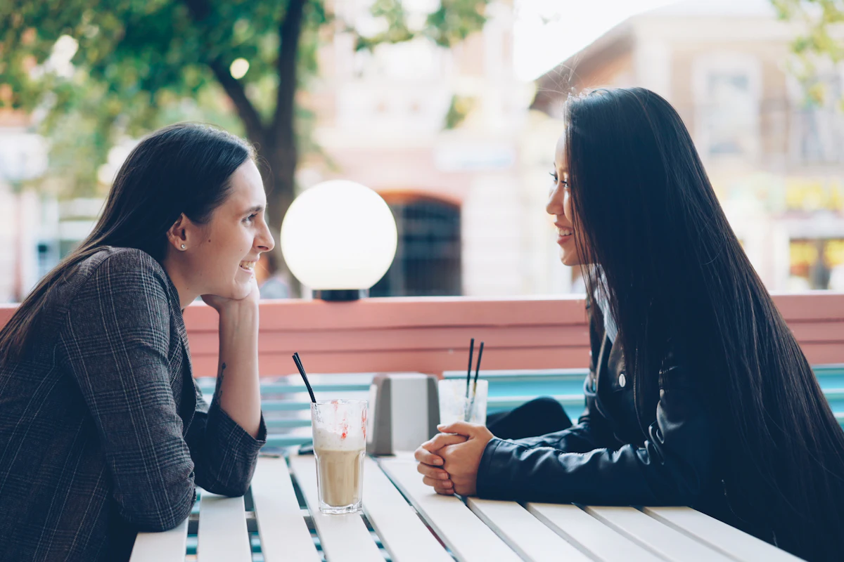 Two women talking face to face across an outdoor cafe table with iced coffees