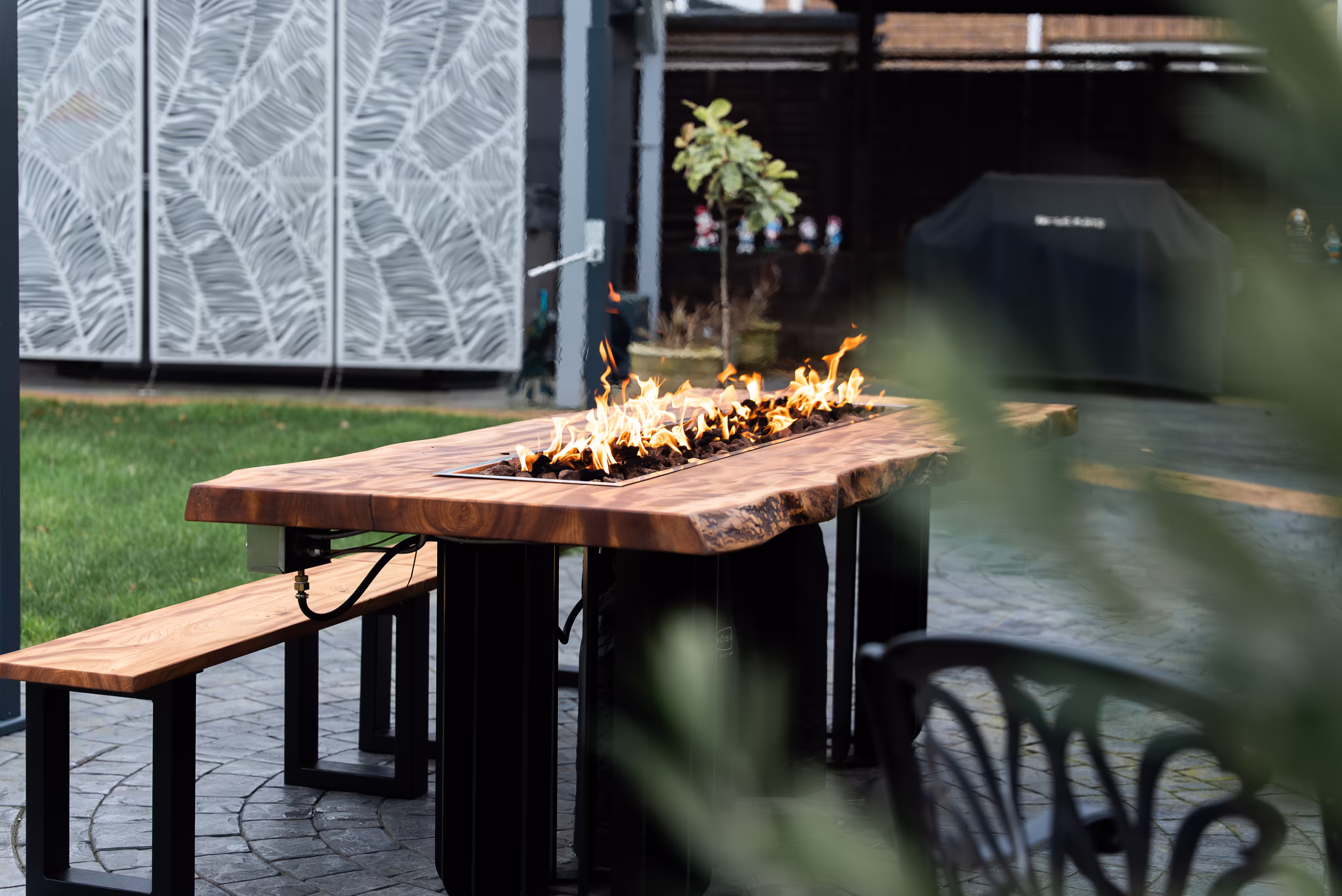 Outdoor rectangular wooden fire pit table with flames, accompanied by wooden benches on a paved patio.