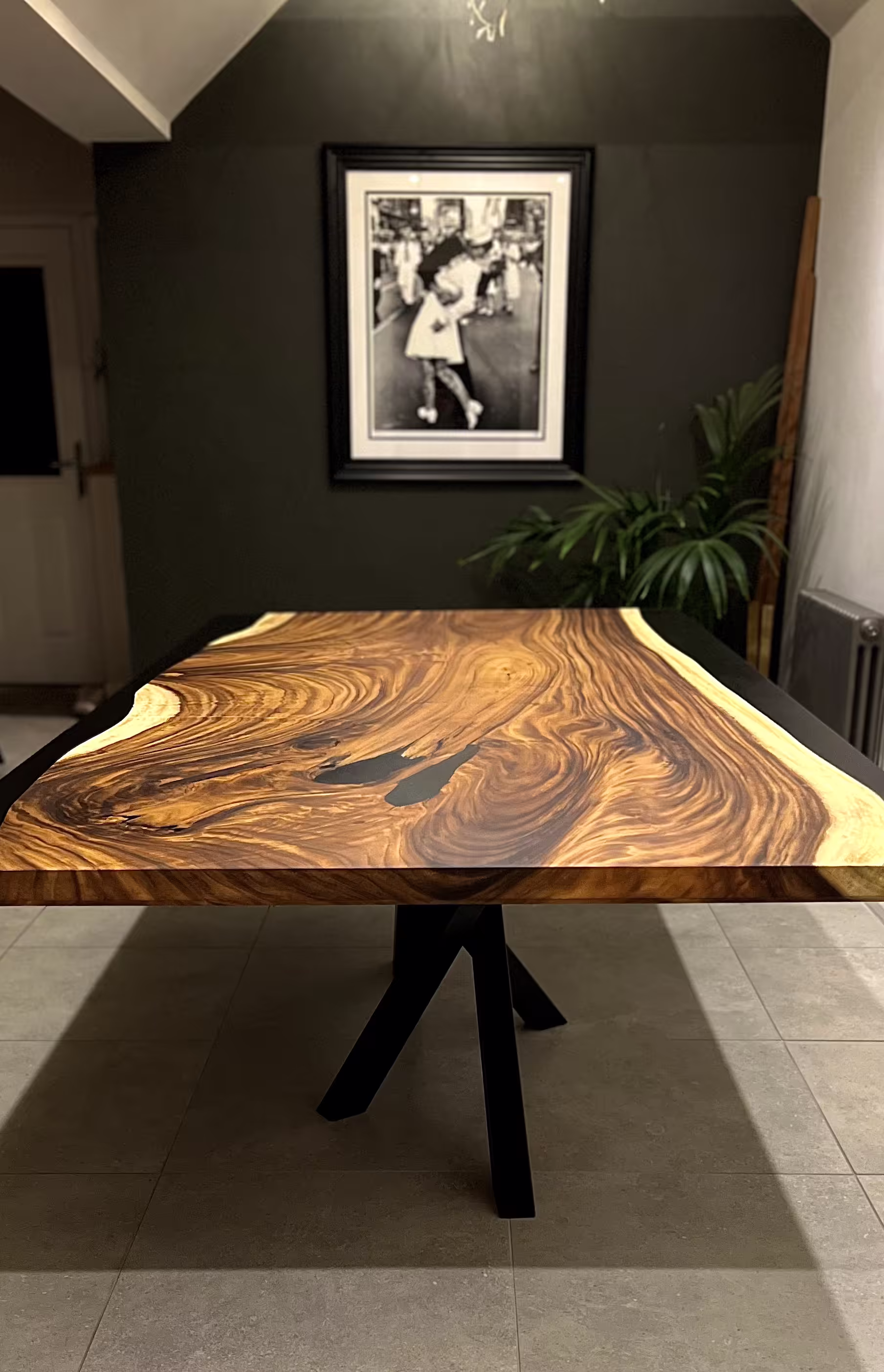 Polished wooden dining table with black epoxy inlays in a modern room with dark walls and a framed black-and-white photo.