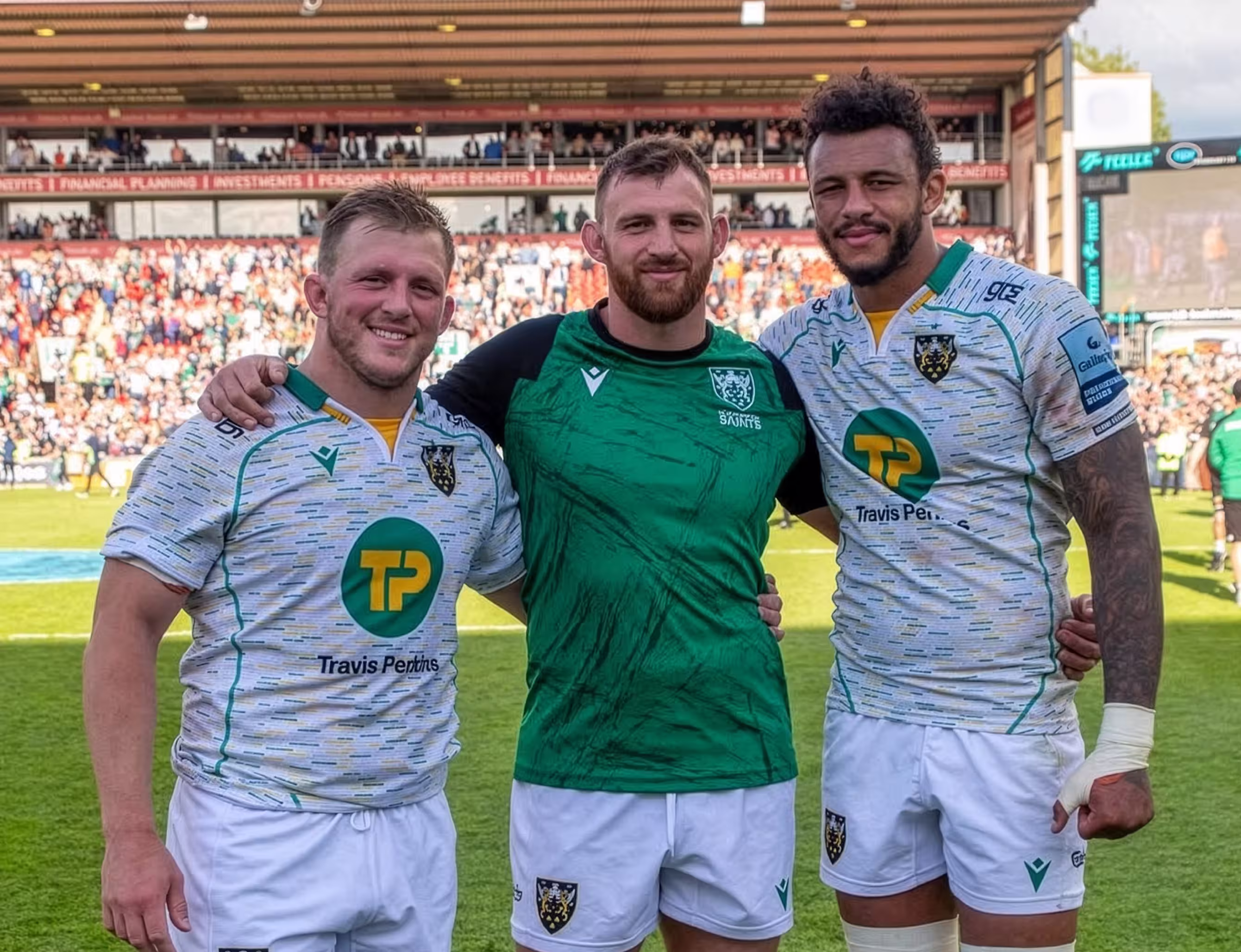 Three rugby players standing on a field with arms around each other, two in white jerseys and one in a green jersey, with a stadium crowd in the background.