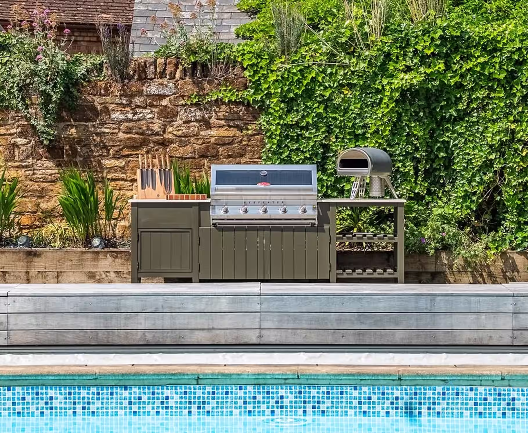 Outdoor kitchen with stainless steel grill and knife set next to a swimming pool, with a stone wall and greenery in the background.