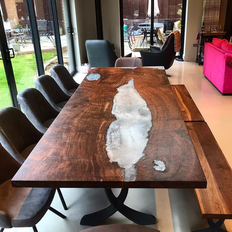Modern dining table with a polished wood surface featuring a white resin inlay, surrounded by grey chairs and a wooden bench in a bright room with glass doors.