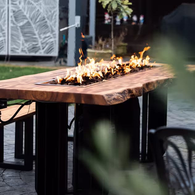 Outdoor wooden table with live fire pit in the center surrounded by benches on a patio.