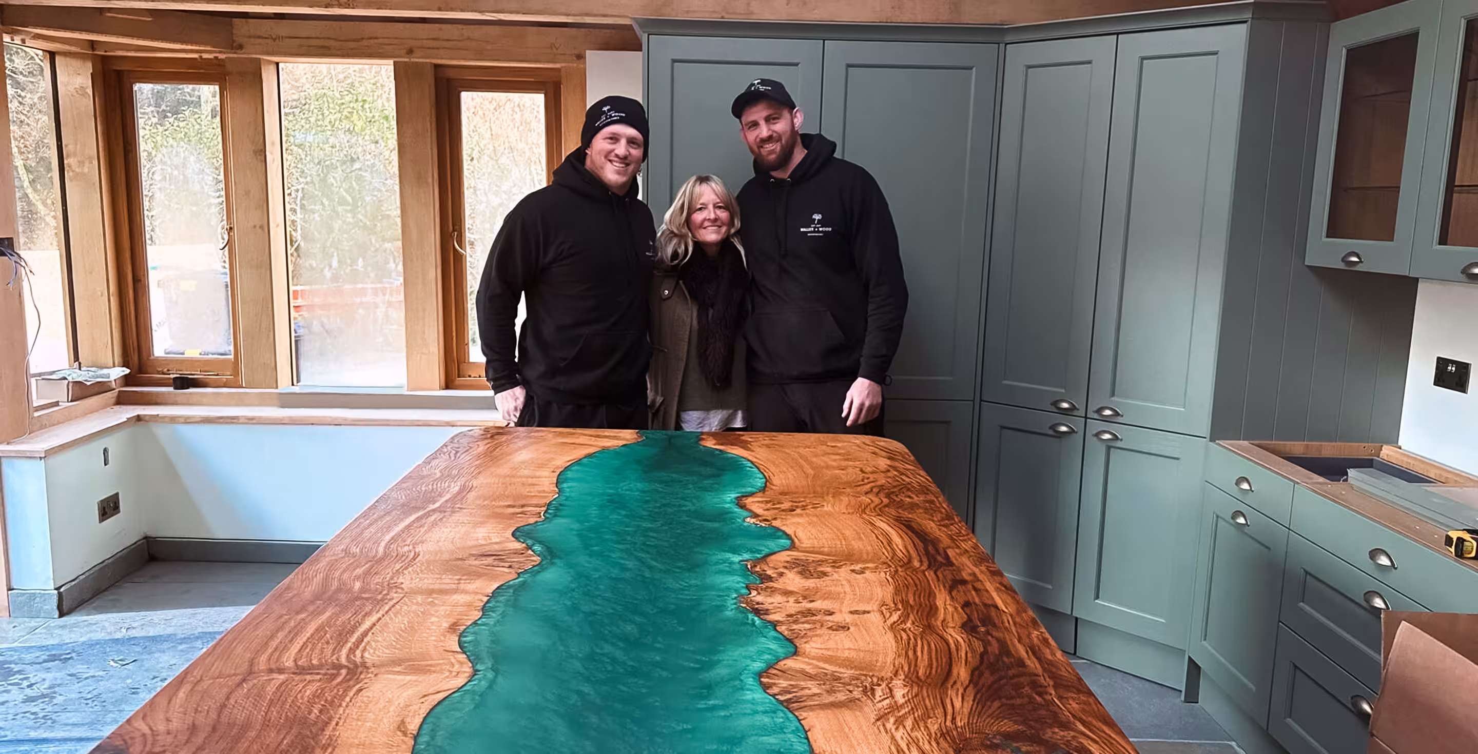 Three people smiling behind a large wooden table with a central blue resin river in a kitchen with green cabinets.