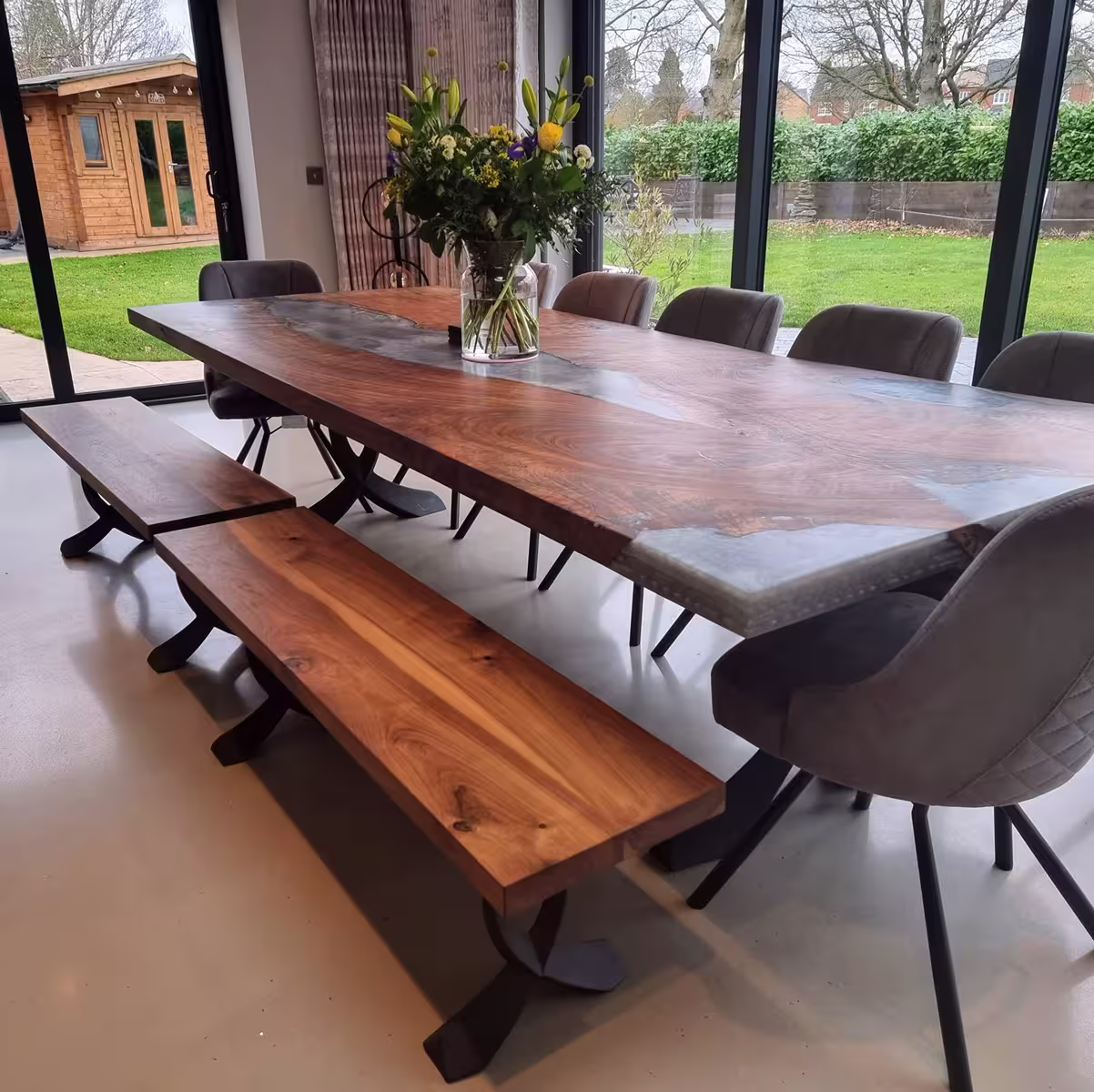 Modern dining table with polished wood and resin finish, surrounded by gray chairs and wooden benches, with a vase of flowers on top and large windows showing a green garden outside.