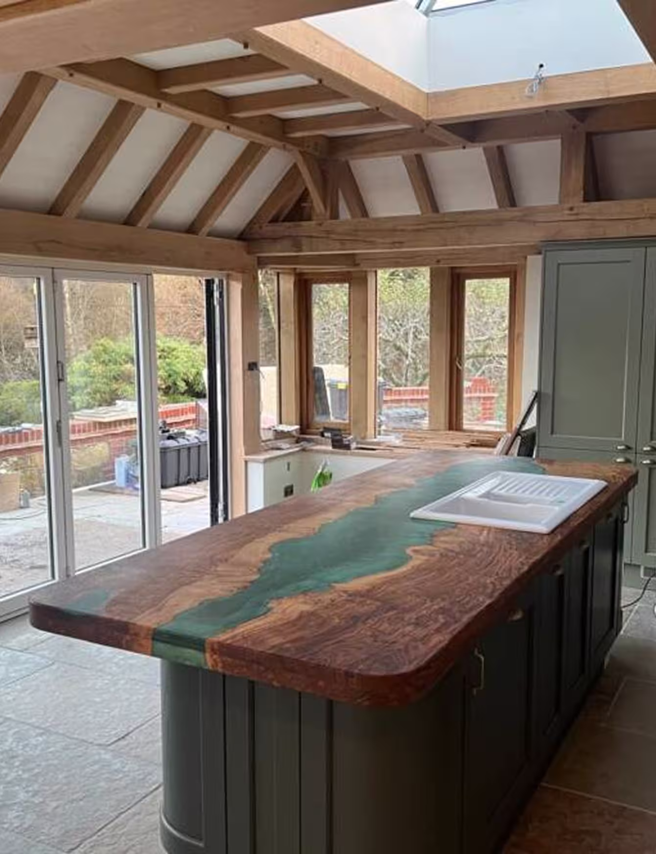 Kitchen interior with a large wooden island featuring a green resin river design and a white sink, under a skylight and wooden beams, with glass doors and windows overlooking a garden.