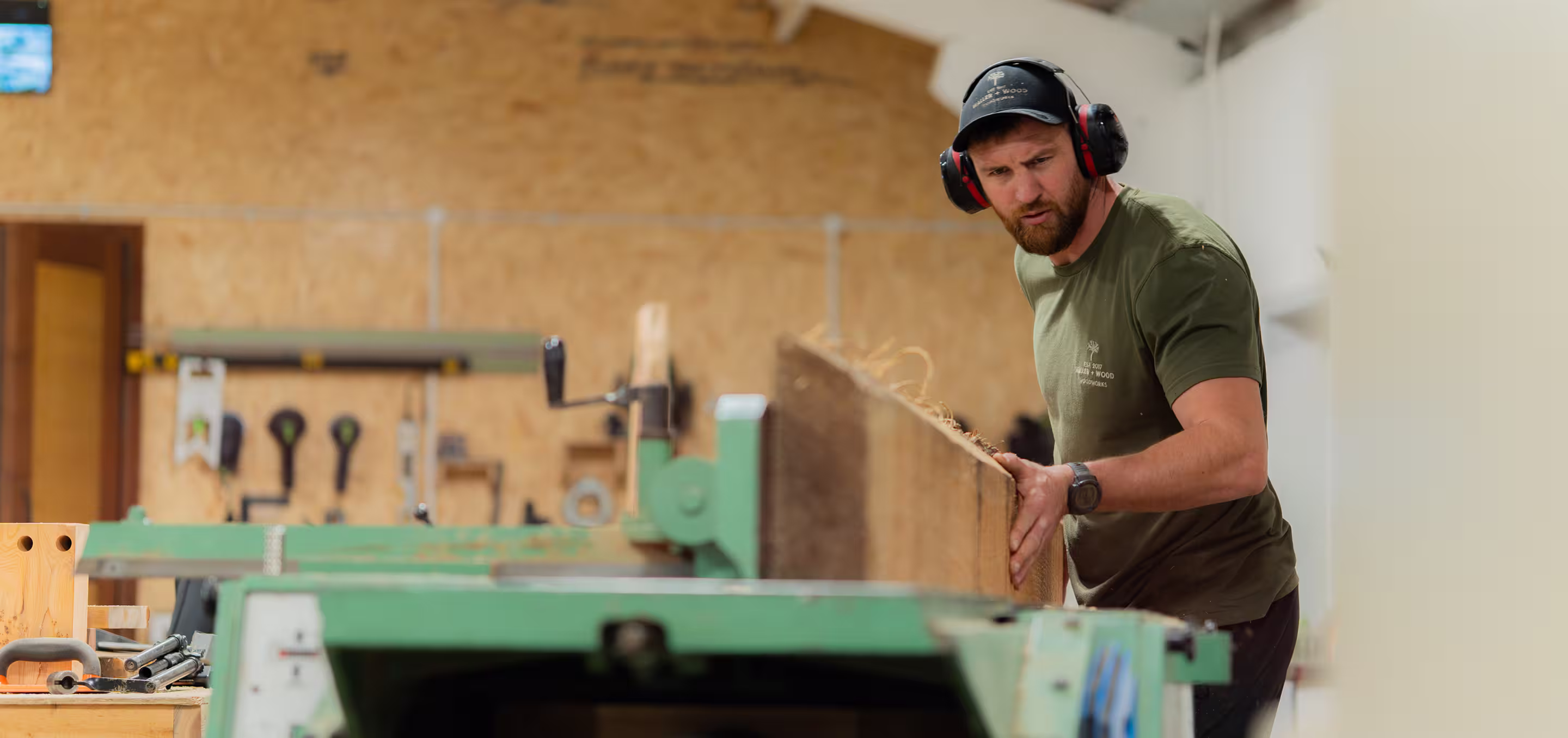 Tom Wood wearing ear protection and a cap planes a wooden board in a workshop.