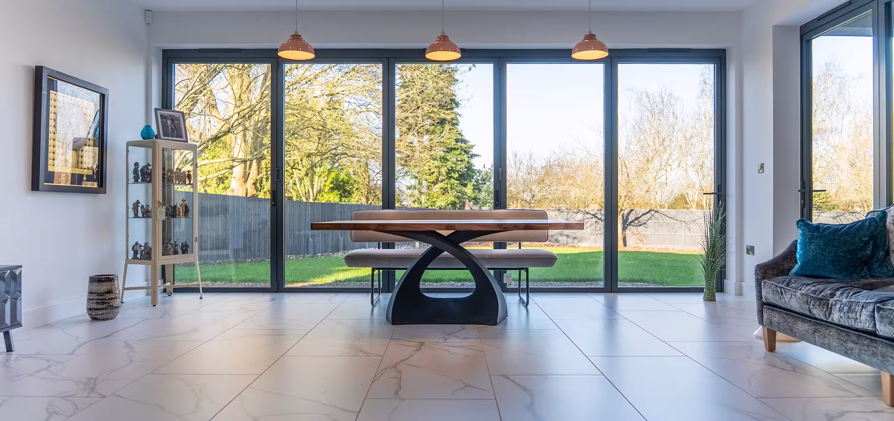 Modern dining room with large glass sliding doors, a wooden table with unique curved black base, bench seating, and bright outdoor garden view.