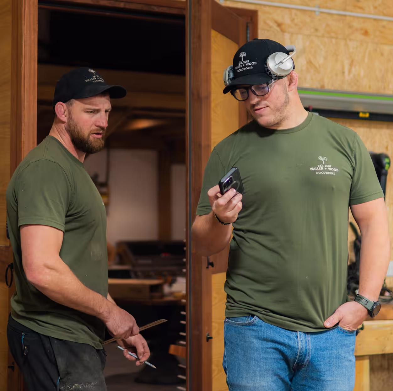Tom and Alex in green shirts and black caps working in the workshop, one holding a pencil and the other holding a smartphone.