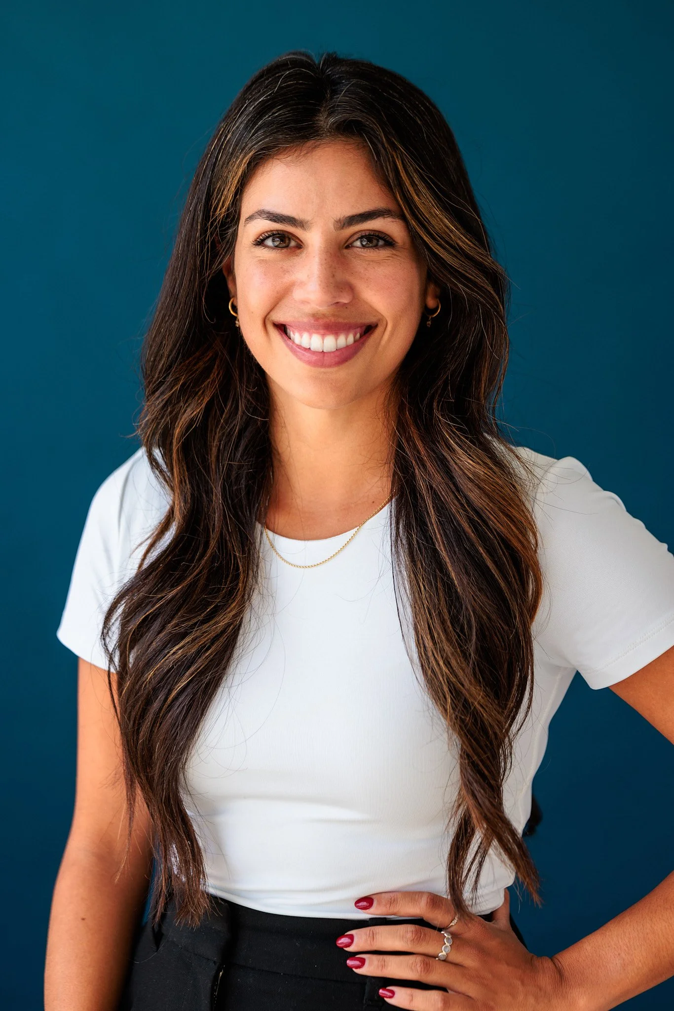 Smiling woman with long dark hair, wearing a white shirt and black pants, standing against a blue background.