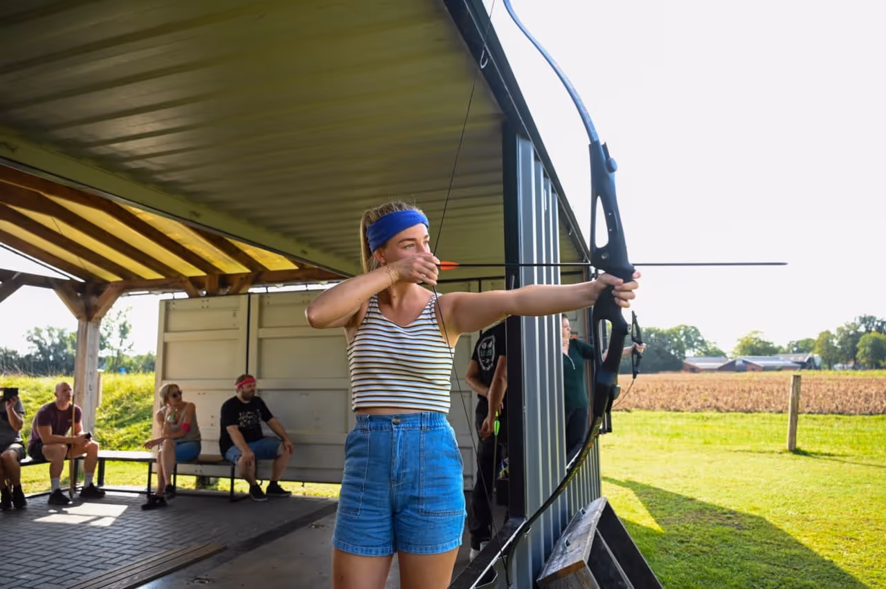 Vrouw met een blauw hoofdband die op een boog schiet bij Expedition Outdoor met toeschouwers op de achtergrond.