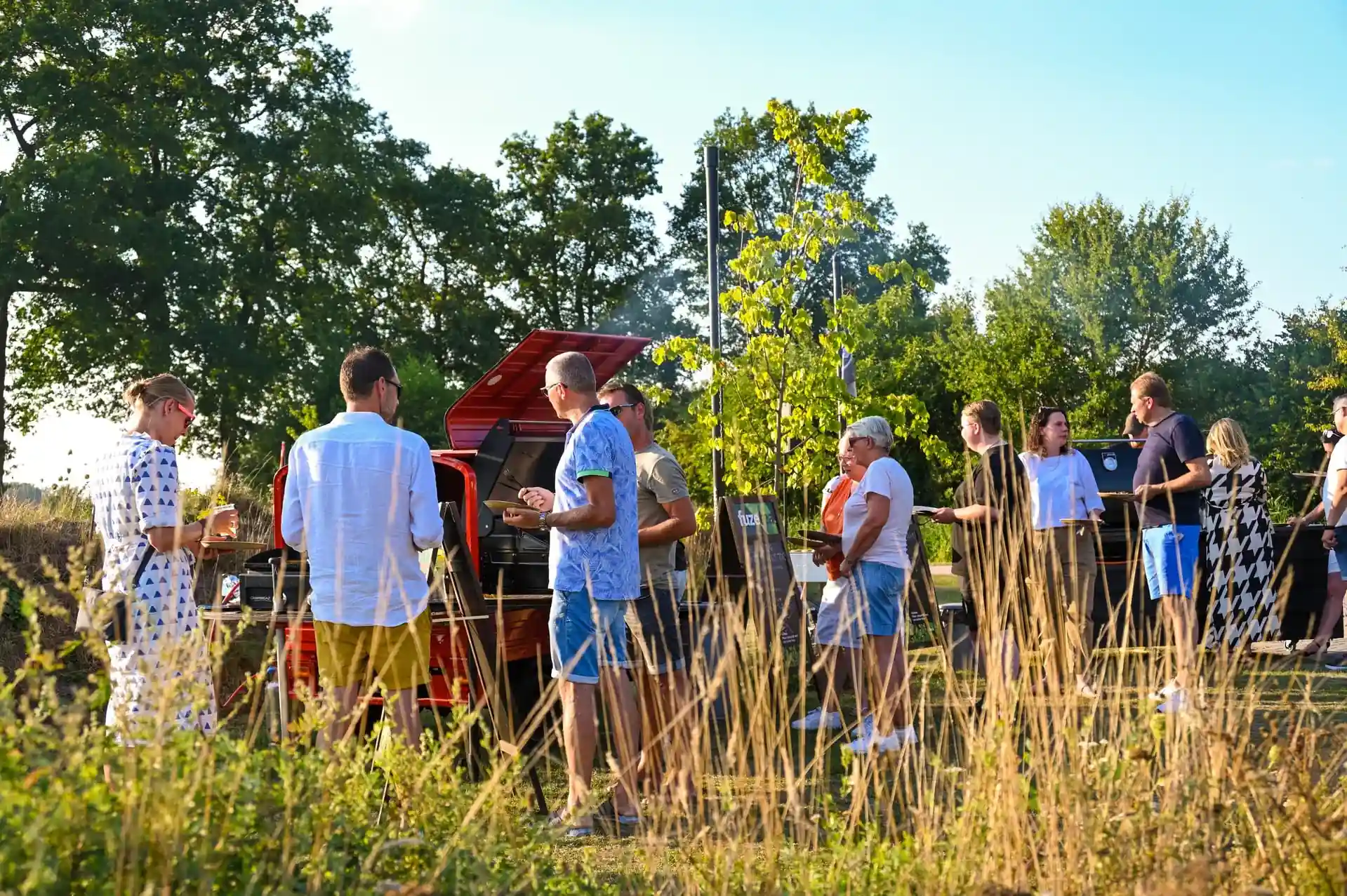 Groep mensen wacht in een rij buiten bij een mobiele eetkraam in een groene parkachtige omgeving op een zonnige dag.
