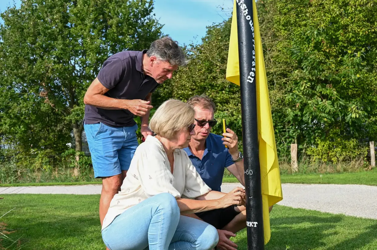 Drie volwassenen onderzoeken een spel dat vast zit aan een zwarte paal in een groene buitenomgeving.