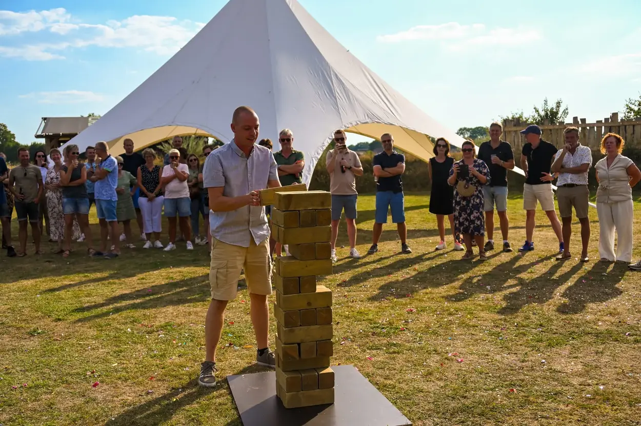 Man speelt gigantisch Jenga-spel buiten terwijl een groep toeschouwers toekijkt bij zonnig weer.