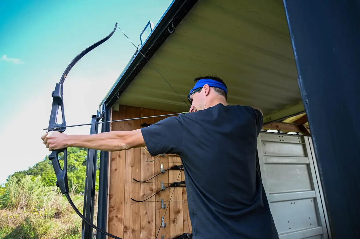 Man met een blauwe hoofdband oefent boogschieten in een schietstand buiten.
