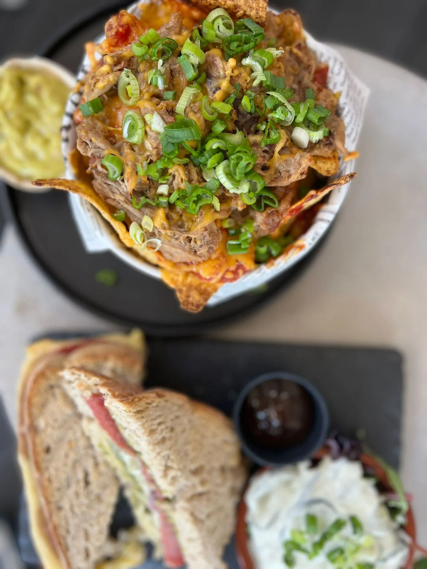 Nacho's met gesmolten kaas, vlees en verse groene ui, geserveerd met een sandwich en een salade met dressing op een zwarte plaat.