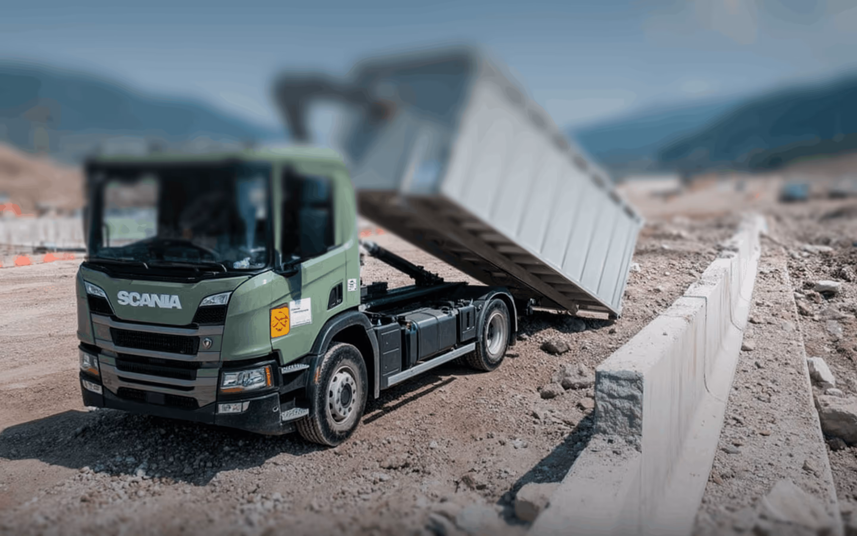 Camion benne vert militaire de marque Scania déversant des matériaux sur un chantier de construction montagneux.