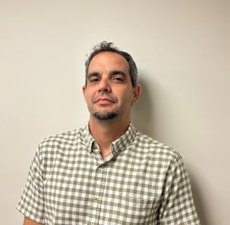 Man with short dark hair and goatee wearing a green and white checkered shirt against a plain beige wall.