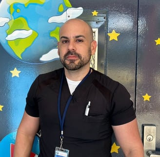 Man with a bald head and beard wearing black scrubs standing in front of a wall painted with a globe and yellow stars.