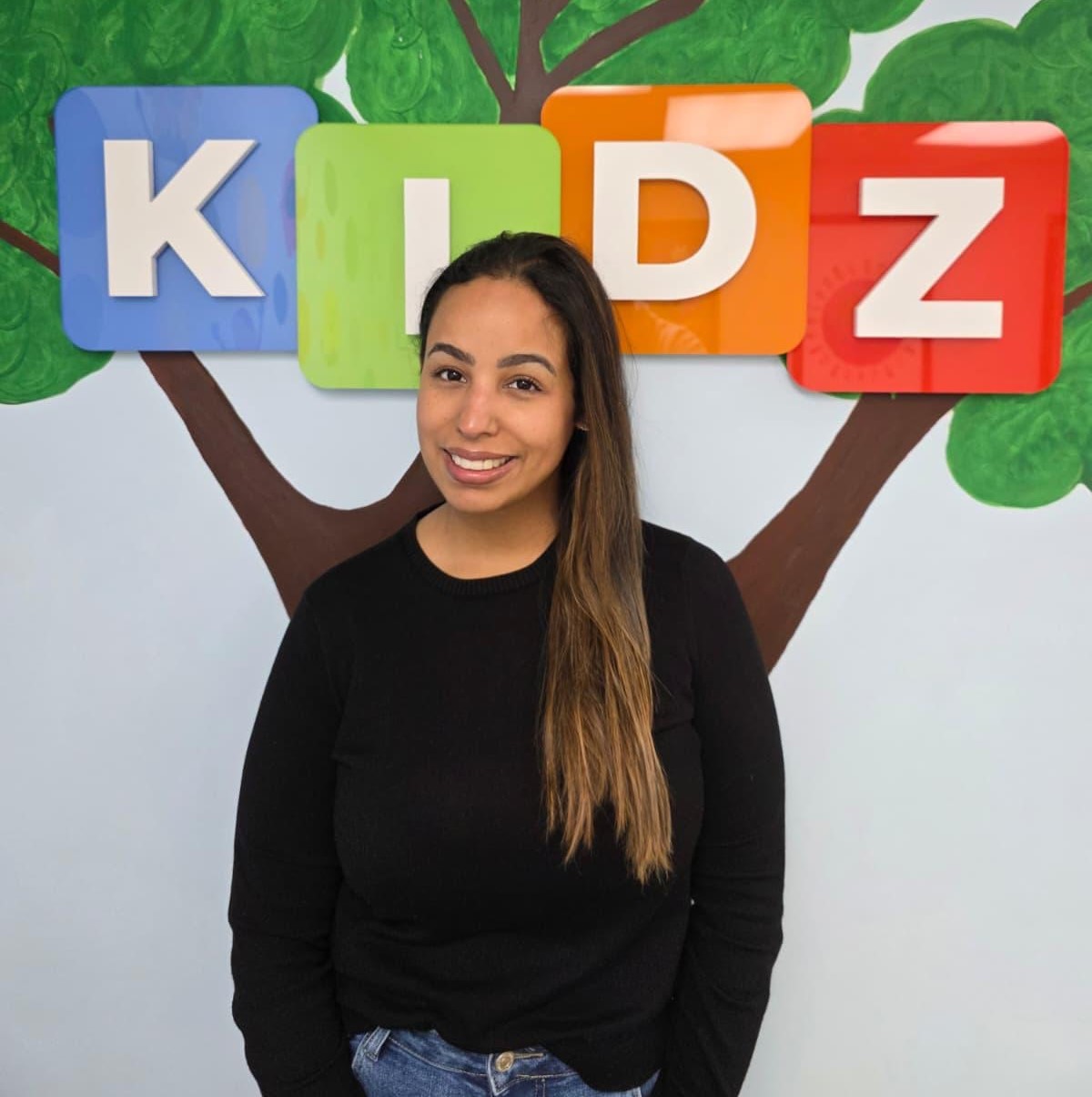 Smiling woman with long hair wearing a black sweater and jeans standing in front of a wall with a painted tree and colorful blocks spelling 'KIDZ'.