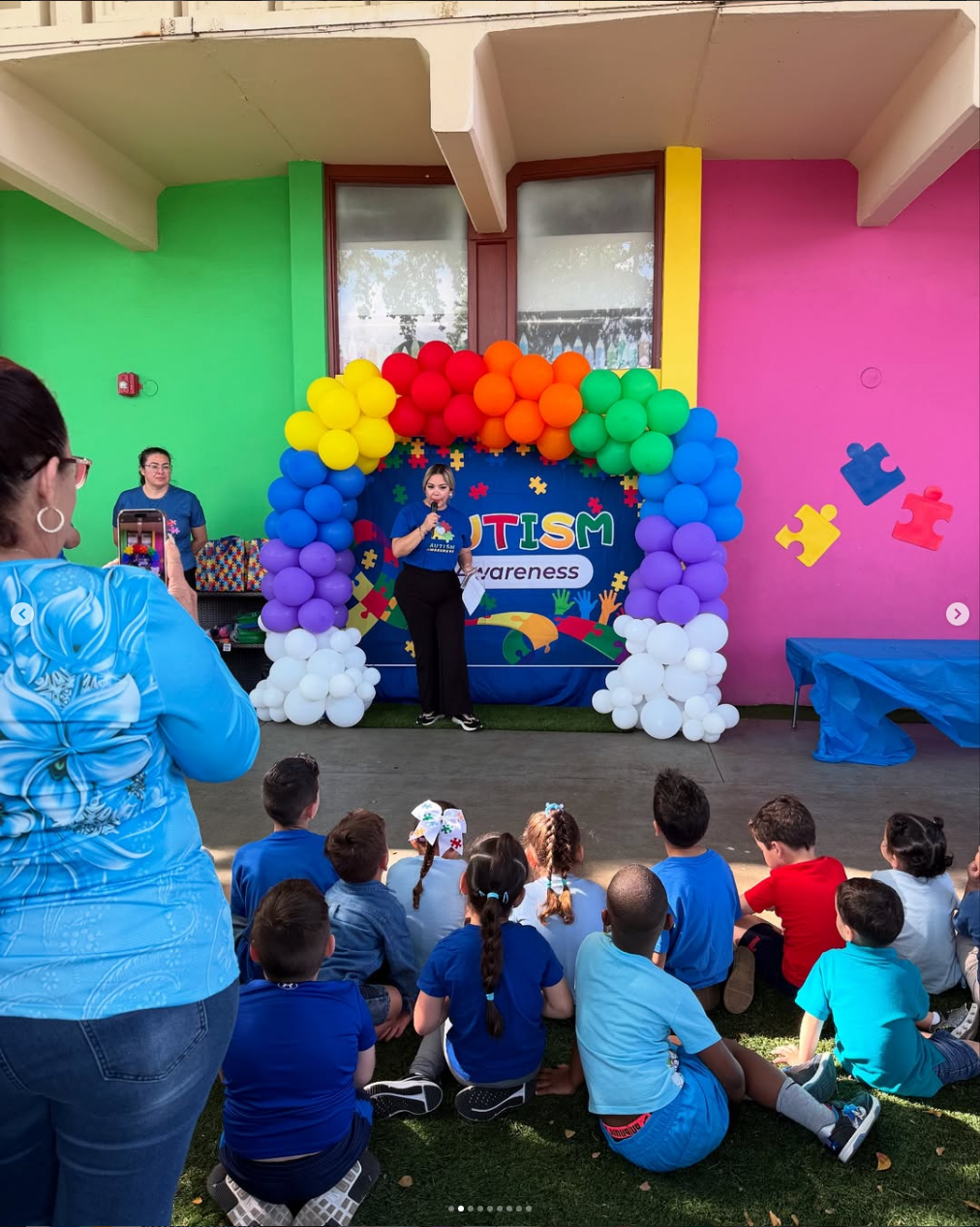 A woman speaking with a microphone under a rainbow balloon arch at an Autism Awareness event, with children seated on the ground watching.
