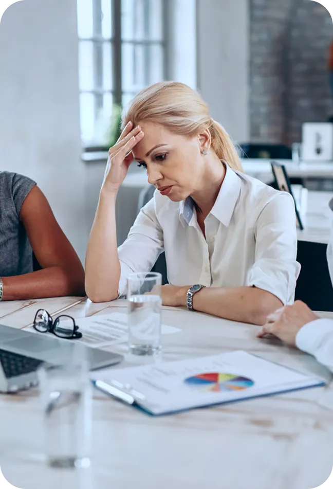 Stressed woman with blonde hair sitting at a table with documents and a laptop, holding her forehead in an office setting.