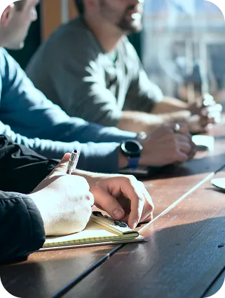 People sitting at a table writing notes in a notebook during a meeting.